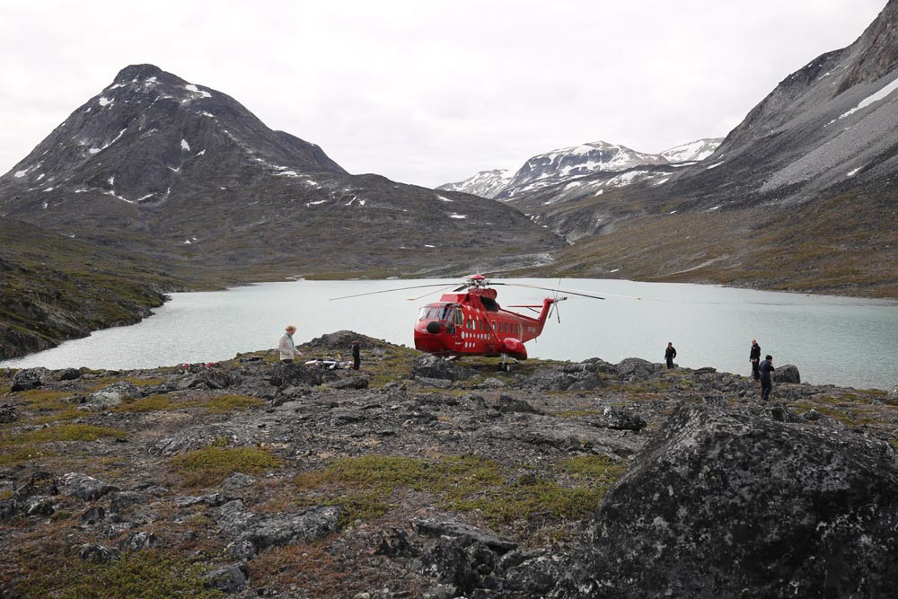 "A red heliocopter lands by a body of water in Greenland, Greenland’s glacier retreat rate has doubled over past two decades"