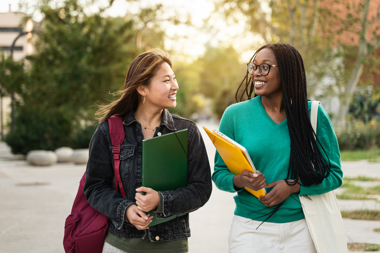 Two smiling students walking on a campus with books in hand. 