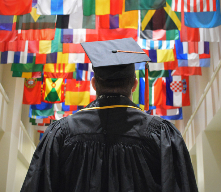 Student in graduation cap and gown standing in front of international flags. 