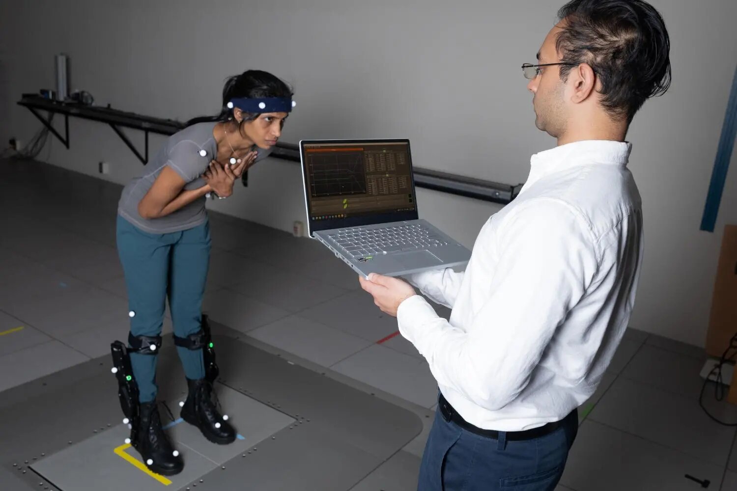 To Help Recover Balance, Robotic Exoskeletons Have to be Faster Than Human Reflexes; "Postdoctoral fellow Surabhi Simha demonstrates how researchers tested whether robotic ankle exoskeleton boots can help people with their balance when its disrupted. Subjects wore sensors for motion capture, muscle activity, and ultrasound while they stood on a platform that shifts abruptly to disrupt their balance. Research engineer Rish Rastogi, right, monitors some of the data being collected."