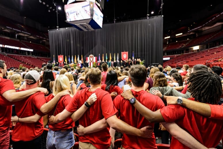 Students in Red T-shirts linking arms in front of a stage with flags on it.