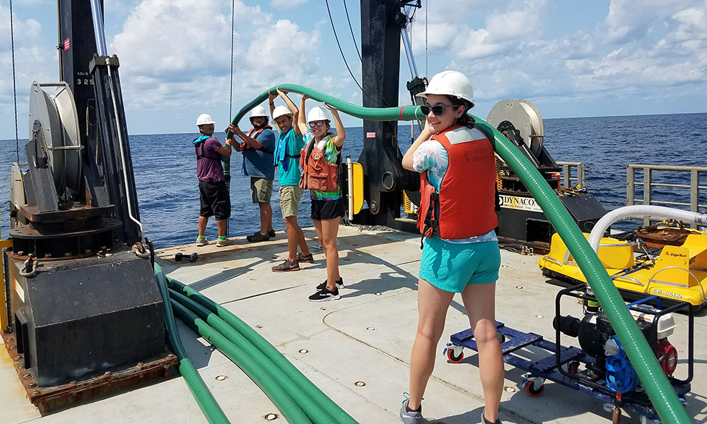 Five college students wearing hard hats and life vests holding a green pipe above their heads while on a barge in the ocean