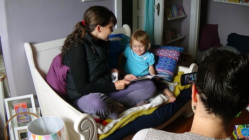 A woman reads to a child while another woman records it on a cell phone.