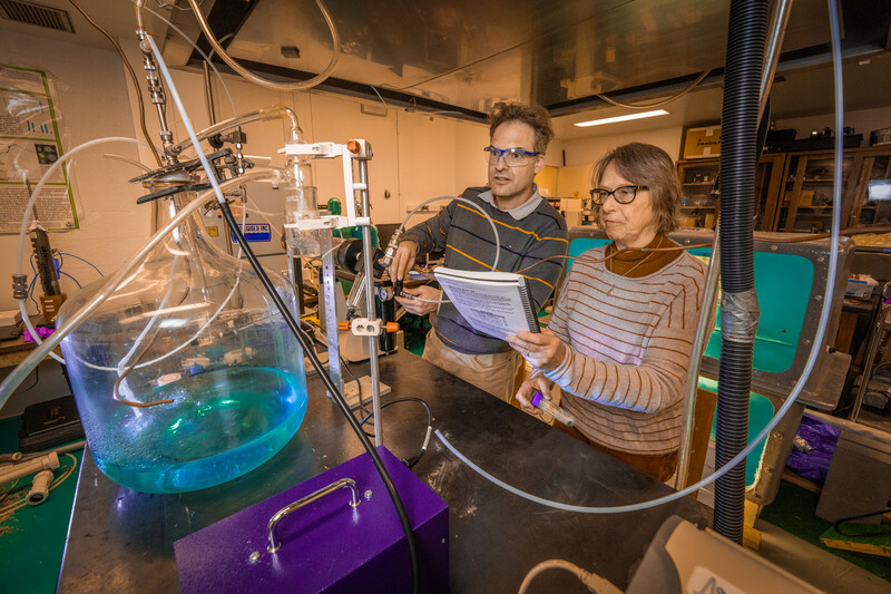 Two scientists standing in a lab, working on an experiment with a large jar of a blue solution and a lot of plastic tubing.