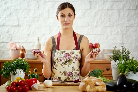 A woman in an apron holds up an onion and garlic bulb.