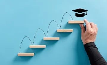 A hand arranges wooden blocks in ascending order, placing a graduate cap on the highest rung, to demonstrate the achievement of college graduates.