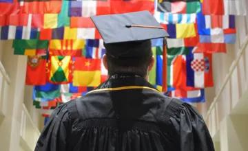 Student in graduation cap and gown standing in front of international flags. 