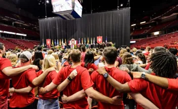 Students in Red T-shirts linking arms in front of a stage with flags on it.