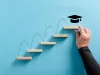 A hand arranges wooden blocks in ascending order, placing a graduate cap on the highest rung, to demonstrate the achievement of college graduates.