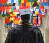 Student in graduation cap and gown standing in front of international flags. 