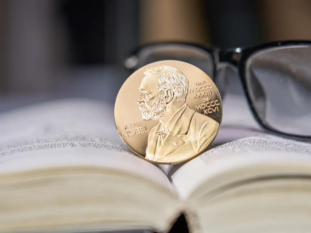 Nobel Prize coin sitting atop an open book beside black framed glasses.
